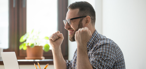 digital-collaboration.png Joyful bearded man raising fists in triumph in front of a laptop computer at a brightly lit home office.