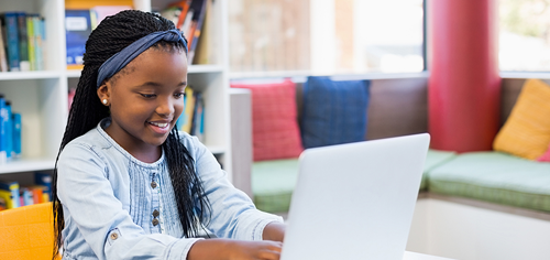 virtual-whiteboard-and-chat.png Young girl smiling while using a laptop in a colorful library setting.