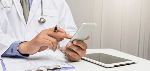 Medical professional in a lab coat using a smartphone with a stylus, clipboard and tablet on the desk.