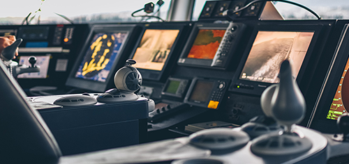 Bridge of a ship with navigation equipment including multiple screens and steering controls.