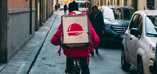 Get inspired rideshare Delivery person in a red jacket with a food delivery backpack cycling on an urban street.